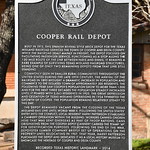 Cooper Rail Depot (Cooper, Texas) Historical marker for the old Texas Midland Railroad depot in Delta County, Texas.  The plaque states:

Cooper Rail Depot - Built in 1913, this Spanish Revival style brick depot for the Texas Midland Railroad serviced the town of Cooper and Delta County. While the railroad dealt mainly in freight, the depot focused on facilitating passenger service, functioning as a stop along the 130-mile route of the line between Paris and Ennis. It remains a rare example of surviving Texas Midland Railroad structures, being one of only two remaining depots from that line still standing. 

Commonly seen in smaller rural communities throughout the United States during the late 19th century, the arrival of the railroad in 1895 brought a dramatic increase in population and commerce to the isolated town of approximately 300 residents. The following year saw Cooper&#039;s population grow to more than 1,000 and for the next three decades the population steadily increased until it peaked with 2,563 residents in 1925. The Great Depression and cessation of Texas Midland operations in 1934 thwarted the growth of Cooper. The population remains relatively steady to this day. 

The depot remained vacant from the closing of the Texas Midland rail line until World War II. Following the United States&#039; entry into the conflict, local resident Harry Patterson established a cannery operation within the building. Workers canned chicken here that was sent overseas as part of troop rations. Cans originating from the Cooper depot cannery carried the stamp &#039;4P&#039; for identification purposes. Following the end of the war, Depoyster Lumber Company briefly set up operations on the property until relocating in 1967. That year, Harry Patterson purchased the depot and dedicated it as a local history museum to showcase the heritage of Cooper and Delta County. Recorded Texas Historic Landmark - 2014