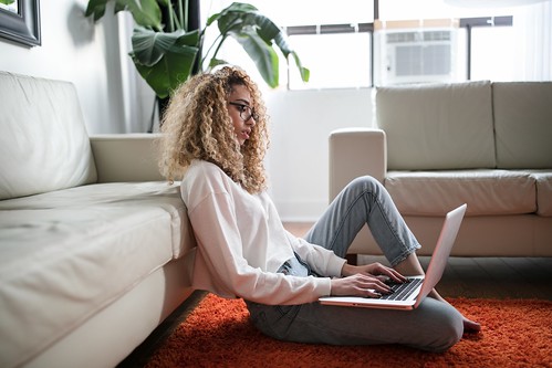 A woman in a white blouse and grey jeans sits on a red shaggy carpet and types on her laptop. She wears black glasses and leans on a white leather sofa - Financial Aid: Common Terms & Definitions