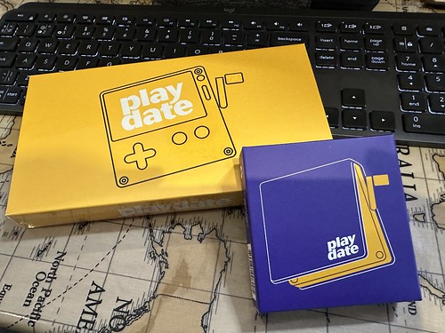 Picture of a yellow rectangular box with a picture of a Playdate handheld and a smaller purple square box showing a Playdate cover, both resting on a table with a world map on it, and inclined on a black keyboard at the top of the picture.