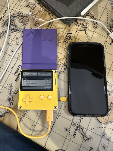 Playdate handheld, purple cover open, showing the Catalog app loading games. It’s connected to USB to charge with an iPhone 11 to the right of it to provide scale.