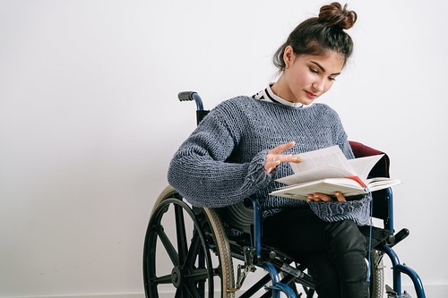 A woman in a wheelchair reads a book. She wears a grey sweater and has her brown hair in a bun - Formas de Solicitar Ayuda con Alojamientos en tu Universidad