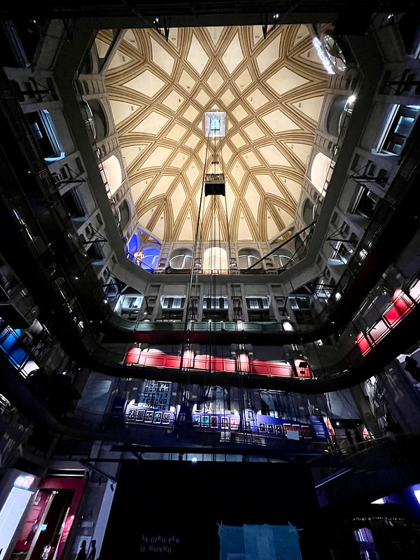 a photo inside a big, dark space, looking up past several levels of galleries around the edge, with a lighter, brighter ceiling. A small dark cuboid of a lift is heading towards a small light rectangle in the centre of the roof.