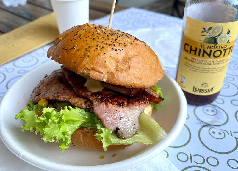 A photo of a seeded burger bun containing several slices of bacon on top of some fresh lettuce. A bottle of Chinotto stands next to it.