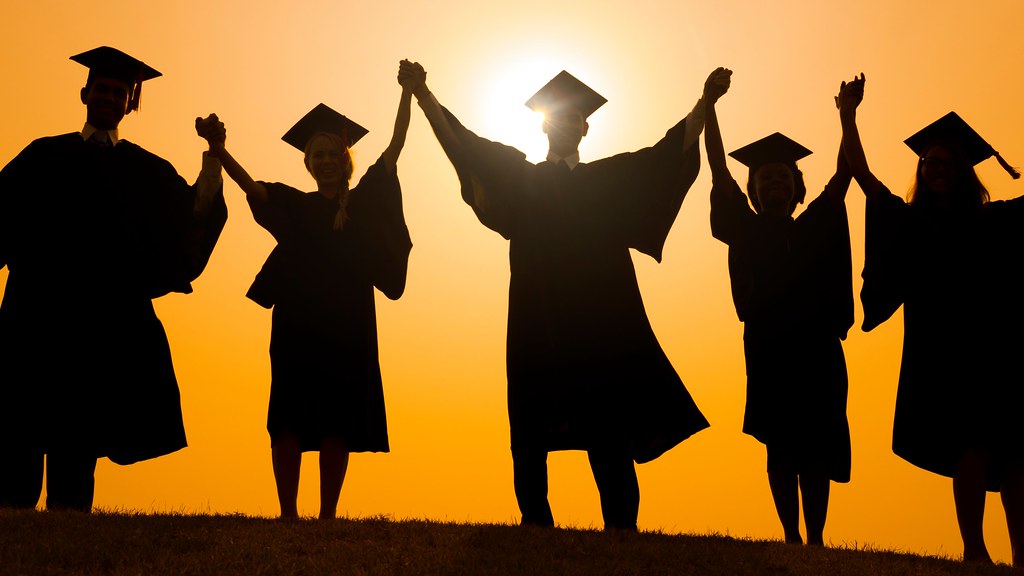 A photo of students in mortarboards in silhouette against the sun raising their arms in triumph