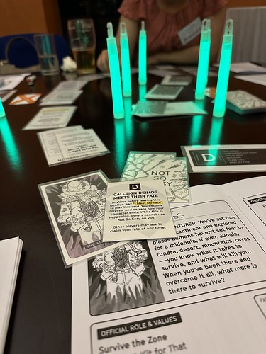 I picture of a game table, in the foreground the adventurer character sheet, in the background, a trail of cards and locations surrounded by green glowsticks