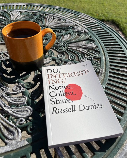 The book 'Do Interesting', on a green metal table next to a mug of black coffee.