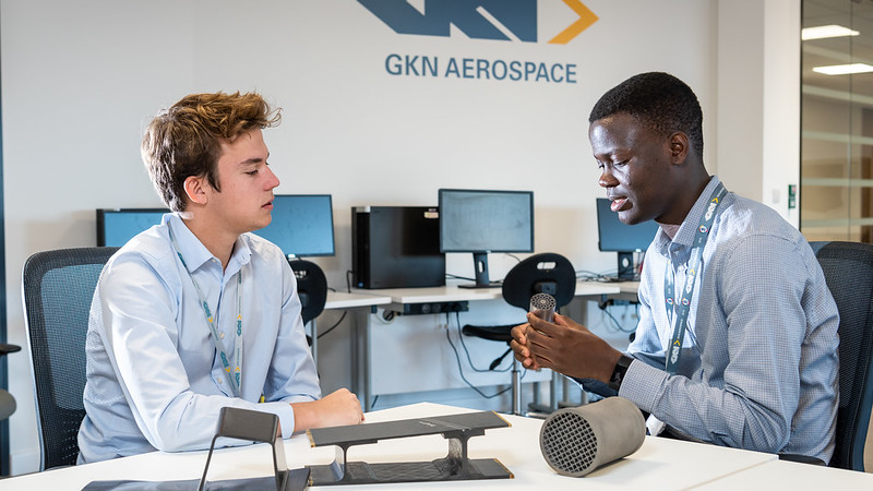 Two young men sit at a table discussing aerospace components before them on a table.