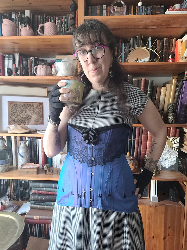 Marion, en corset violet et robe pied de poule, une tasse de thé à la main