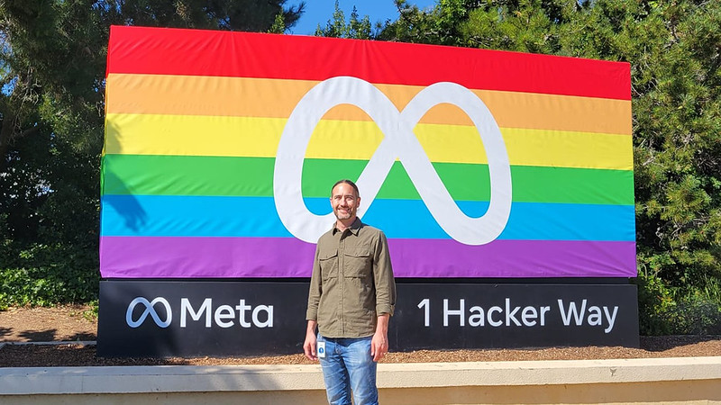 A person (Dr Michael Proulx) stood in front of a Meta sign with a rainbow flag background.