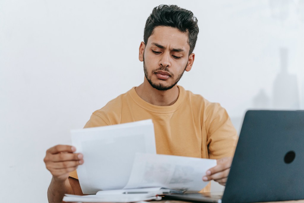 A man wearing a yellow shirt sits at a desk and looks at papers - Use Our First Job Cover Letter Templates! 