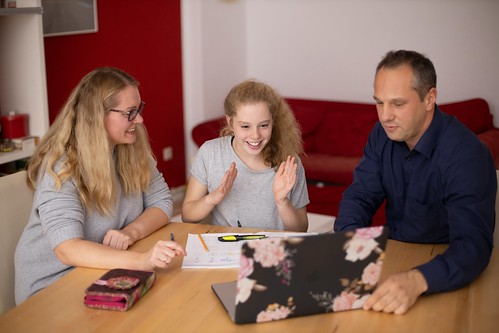 A teenager sits at a table with her parents. The three of them look at a laptop with a floral case. Pens and papers sit on the table - FAFSA Deadlines by State