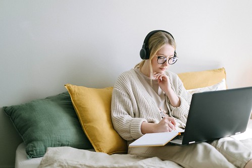 A woman wearing glasses, headphones, and a beige sweater sits on her bed and writes on a notebook that's propped on her laptop - 2024-2025 Better FAFSA Updates 