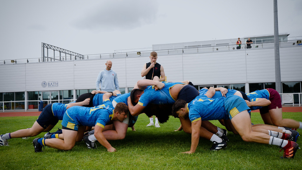 A group of rugby players in blue and yellow University of Bath uniforms locked in a scrum on a rugby pitch.