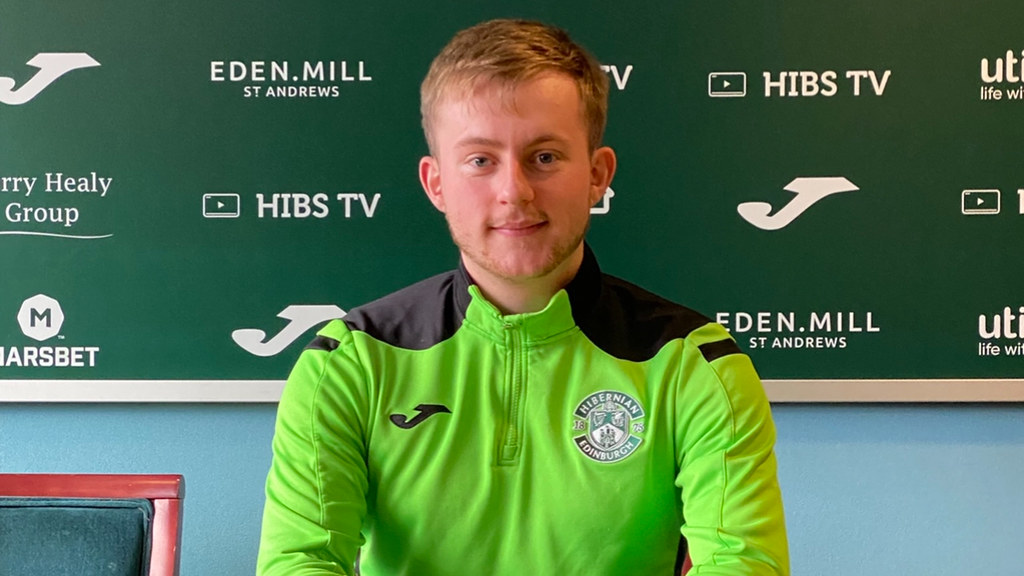 A male student in a bright green Hibernian Football Club branded jumper sits at a table and smiles.