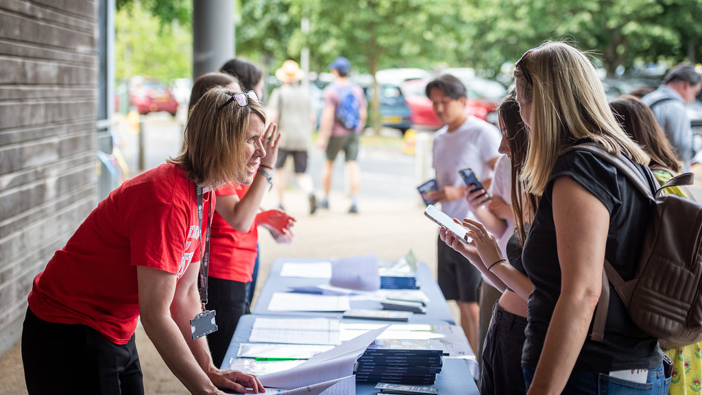 A member of staff in a red University of Bath T-shirt gives advice to some Open Day visitors at an Information Point.