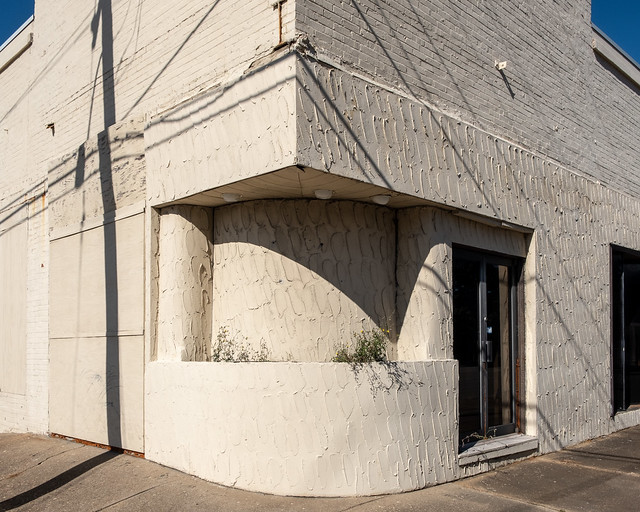 In downtown Newport News, the rounded corner of a commercial building is surfaced in trowel-sweep stucco, and is occupied by a recessed planter of weeds.