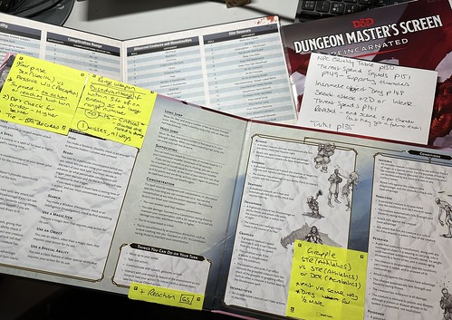 A picture of two GM screens lying on top of each others. The top one is the D&D 5e screen which has a number of yellow post-it notes on with extra rules and table references. Below it, the Acthung! Cthulhu one lies, with a sheet of paper with rules and page reference to be added in on the right. Underneath that it is the outer wrapper from the D&D Reincarnated screen. They all lie on a grey desk.