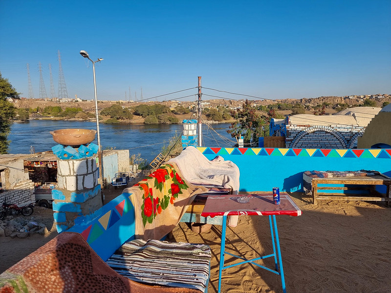 The rooftop of a Nubian guesthouse, with tables and seating areas covered with colourful blankets. In the back you can see River Nile.