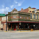 Church Street and Main Street, New Harmony, IN Built in 1910, this “Free Classic”-variant Queen Anne-style building features a one-and-a-half-story section at the street corner with a facade featuring decorative pilasters, large plate glass windows and transoms on the first floor, a chamfered corner, a rooftop balustrade, and decorative panels on the upper portion of the facade, a two-story portion with a buff brick front facade and red brick side and rear facades, a bracketed cornice, an arched parapet, paired one-over-one double-hung windows with transoms, and large plate glass windows and transoms on the first floor, and a large canopy over the sidewalk that wraps around the front of both sections of the building.  The building is a contributing structure in the New Harmony Historic District, designated as a National Historic Landmark in 1965, and listed on the National Register of Historic Places in 1966.