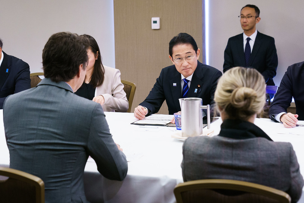 Prime Minister Justin Trudeau sits at a table facing Japanese Prime Minister Kishida Fumio. Other people sitting at the table with them. 
