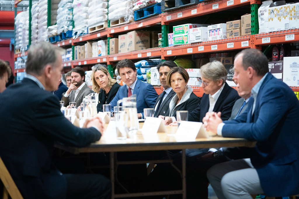 Prime Minister Justin Trudeau sits at a table with Canadian and American stakeholders. Shelves of groceries behind them. 