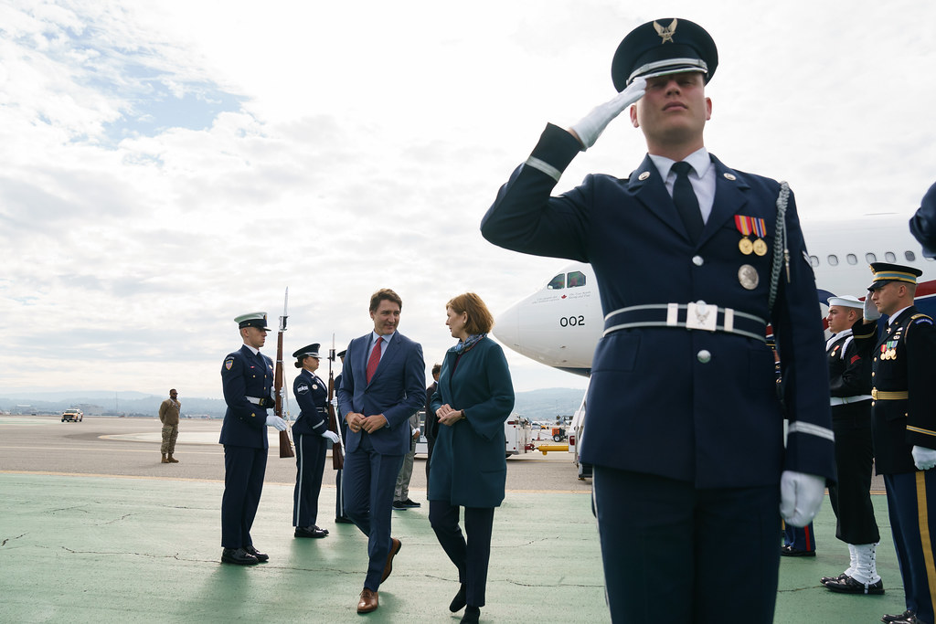 Prime Minister Justin Trudeau walking with a woman on the tarmac. Guards are standing at attention on the tarmac as they walk by. Airplane in the background. 