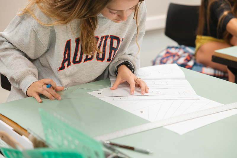 A young woman leans over a desk as she concentrates on drawing straight lines