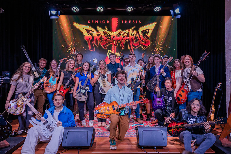 A group of college students stands on stage at a concert venue showing off the guitars they designed