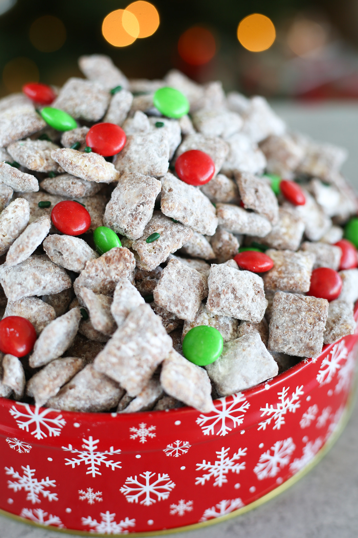 Muddy buddies with red and green M&Ms and green sprinkles in a red snowflake cookie tin