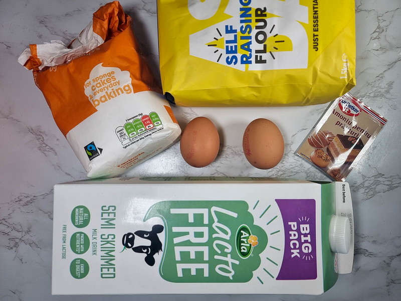 A few of the ingredients for this cake: a green bottle of milk, an orange packet of sugar, a yellow package of flour, two eggs, a small pack of ammonia for cakes