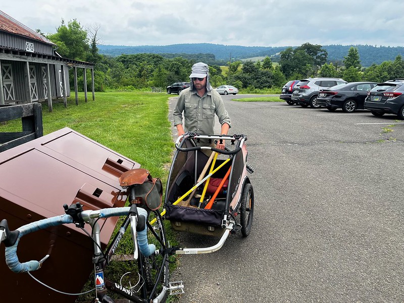 A man standing behind a bicycle trailer full of tools, with the bicycle leaning on a set of bear-proof trash cans in a parking area next to a horse stable barn.