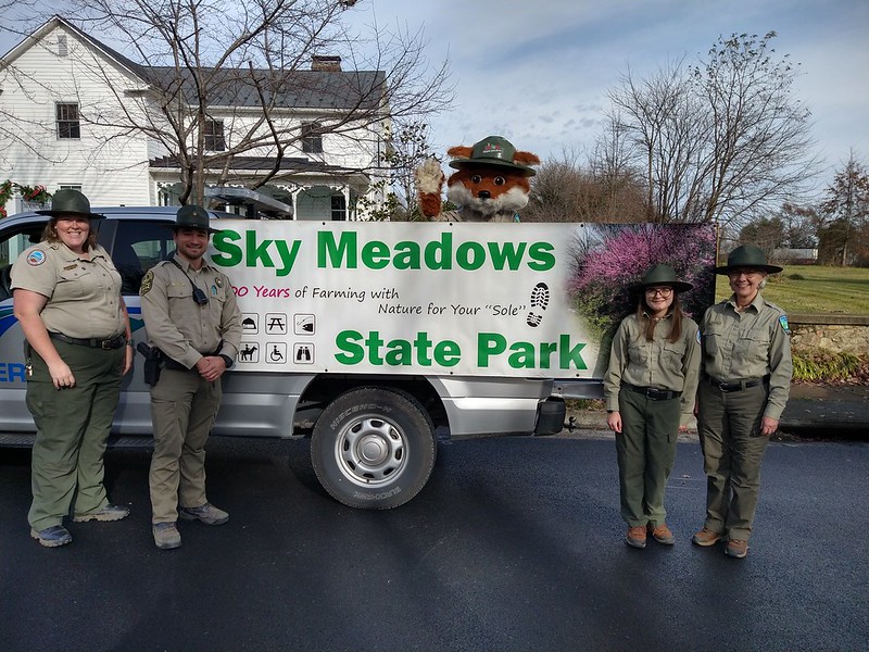 Park rangers and a red fox mascot with a park ranger hat, posing with a park vehicle decorated with a sing that reads "Sky Meadows State Park" 