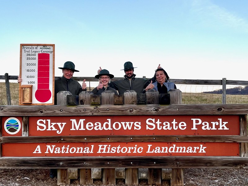 Four smiling park rangers showing thumbs up and presenting a sign that reads "Friends of Sky Meadows Trail Legacy Campaign" with a painted picture of a red thermometer topped up at $28,000