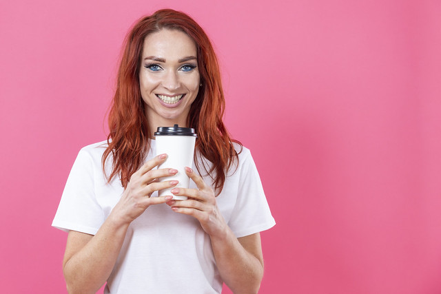 Winsome Female with Ginger Hair Using Disposable Cup of Take Away Coffee, Enjoying Beverage While Wearing White Casual Style Shirt Indoor Studio Shot isolated on pink