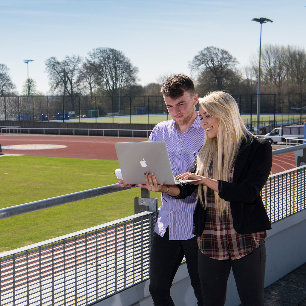 Two young people stood by a running track while using a laptop computer