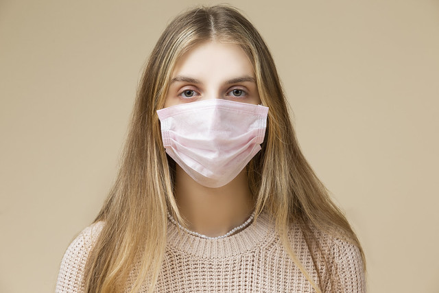 Sick Caucasian Blond Female Girl Wearing Warm Knitted Sweater Using Viral Facial Protective Mask Against Beige.