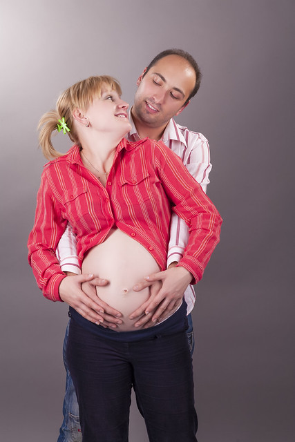 Happy Positive Young Couple Standing Together Close While Hugging HEr Pregnant Woman And Wearing Casual Clothing Over Gray Background