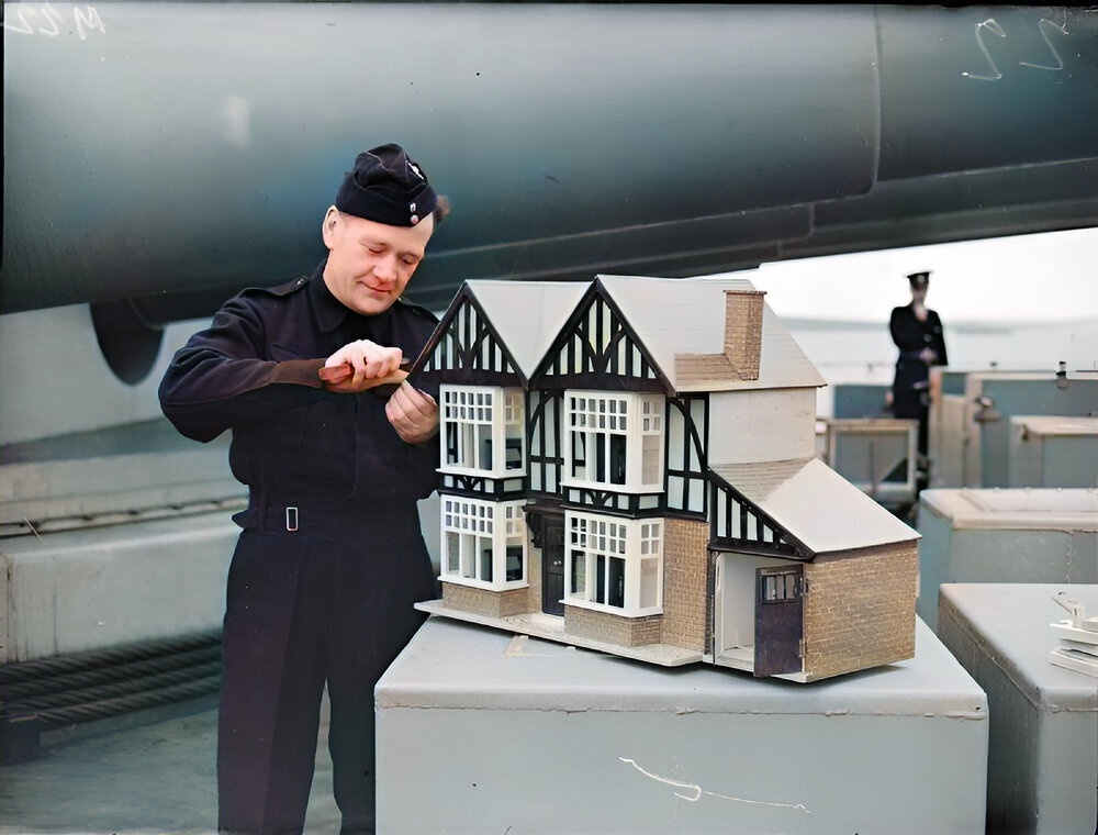 On the foc'sle of a battleship, in the shadow of the guns, a Royal Marine, J Lynch of Newport, Monmouthshire is putting the finishing touches to a large dolls house, complete with furniture, 1943