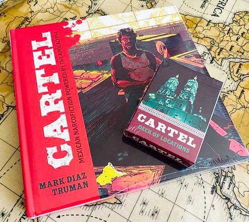 A photograph of a book and a deck of cards. The books is titled 'Cartel - Mexican Nacrofiction Powered by the Apocalypse' and written by Mark Diaz Truman. It has a picture of a man in a blood stained white vest sitting in a backroom with drugs around him. The card deck is titled 'Cartel, Deck of Locatons' and shows a large church.