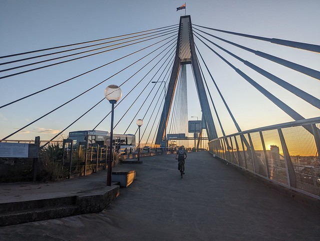 Photograph of a lone cyclist ascending Anzac Bridge cyclepath from the east, in front of the bridge tower and cables and the setting sun