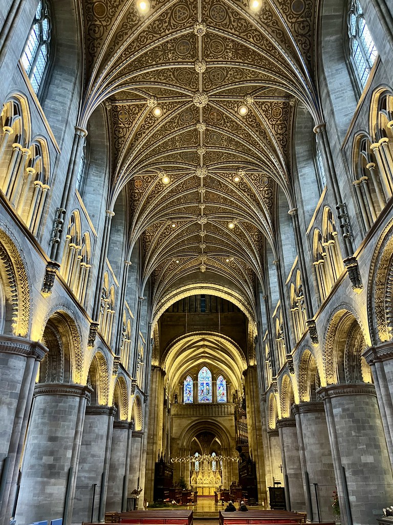 A photo inside a cathedral, the pews just visible at the bottom of the frame, looking up to an illuminated ceiling high above. It's all grey stone, lit with yellow lights, big arches on either side supporting arched windows above.