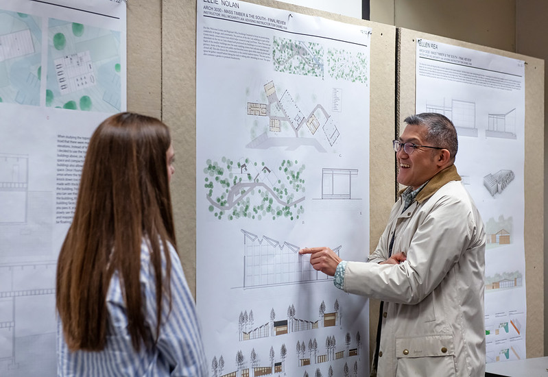 A smiling man speaks with a student as her design drawings hang on the wall behind them.
