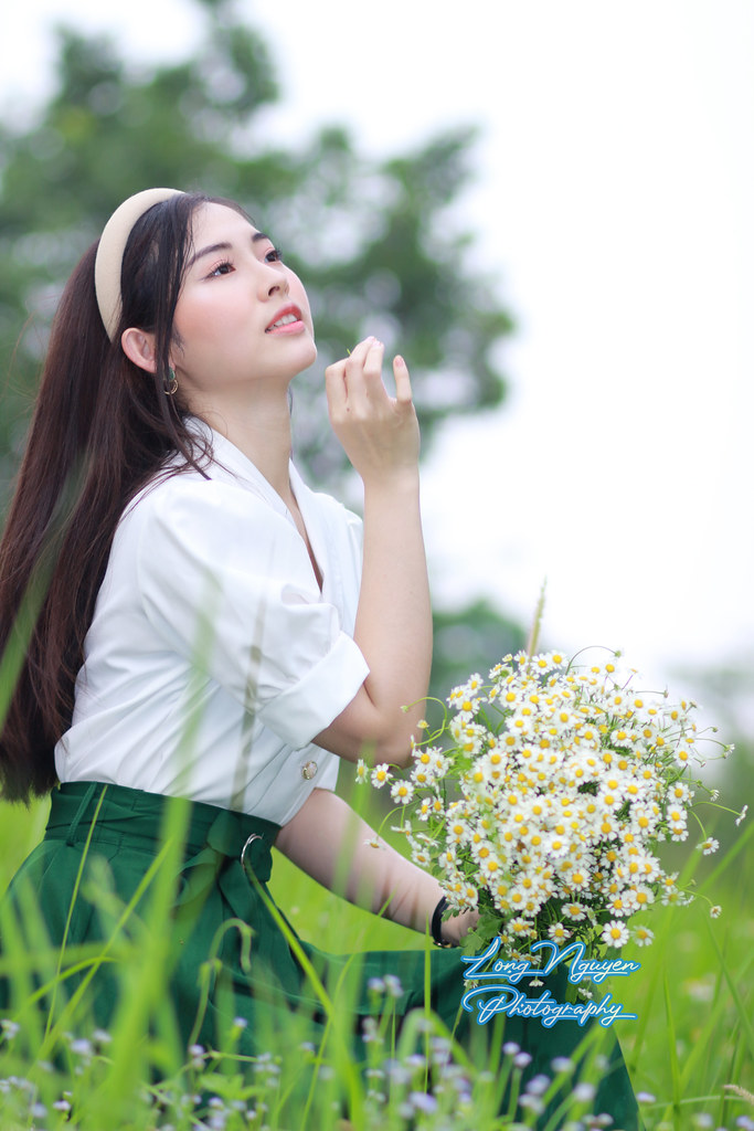 A beautiful girl donning a white shirt, a green waist-belted skirt, and a turban and holding white daisies graces the green lawn with some wildflowers and the chair, her dark hair subtly mixed with red.