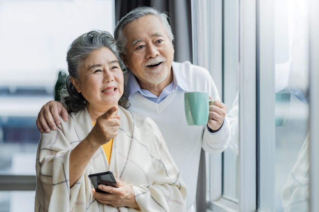 portrait of Asian senior retired couple smiling and looking out of window apartment while hold wife shoulder .Cheerful asian senior couple retirement life. wellness Senior healthy lifestyle concept.