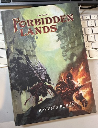 The hardcover roleplaying supplement 'Raven's Purge' lying on a desk and a silver and white Apple keyboard. The cover shows an pale elf-like warrior with a cloak of ravens facing down a burning armoured figure. Text on book: "Free League - Forbidden Lands - Raven's Purge".