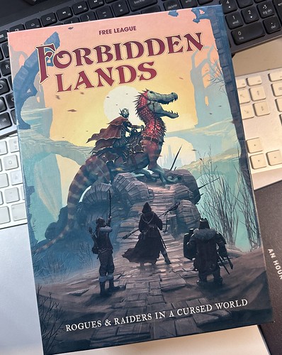 Forbidden Lands box set lying on a desk on top of two keyboard (lower one silver & white Apple, upper gunmetal and black Logi MX). The cover of the box shows ruins with a lizard creature with a rider on stopped on a bridge. The foreground shows three adventurers, one with a drawn bow, looking ready to confront the enemy. Text on box: "Free League - Forbidden Lands - Rogues & Raiders in a Cursed World"