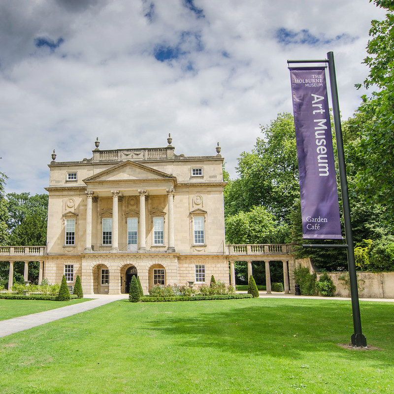 The exterior of The Holburne Museum; a large Georgian building in a garden. A large purple banner is in the foreground displaying the text 'The Holburne Museum Art Museum Garden Café'.