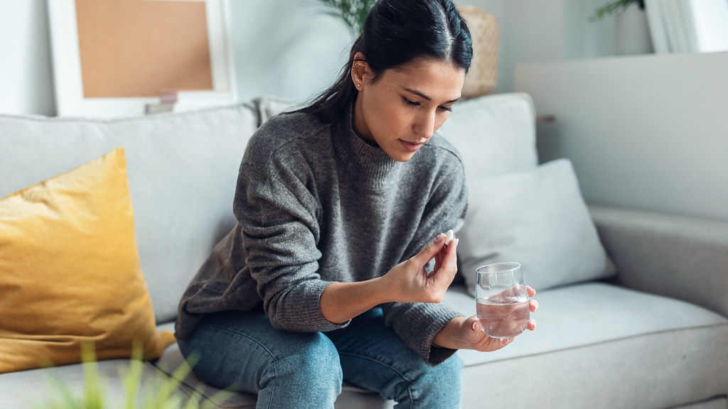 Sad-looking woman sitting on sofa looking at a pill she's about to take with a glass of water