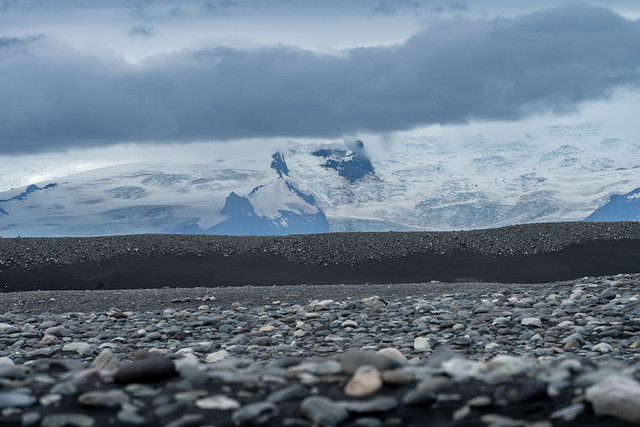 Barren volcanic landscape on a black sand beach, with mountains and glacier in the background. Iceland, south coast