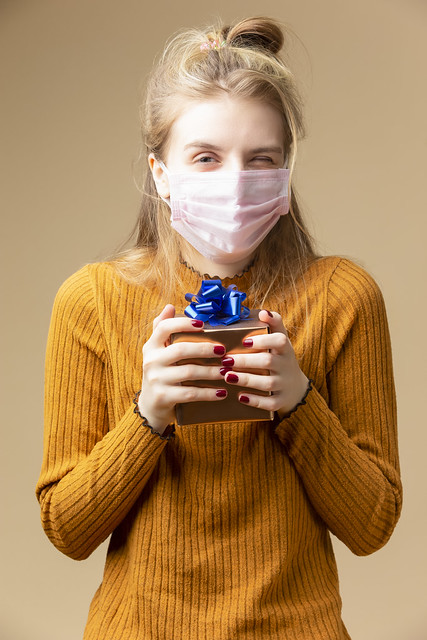Blond Female Girl with Tiny Small Golden Gift Christmas Box Wearing Facial Protective Mask Against Viruses and Unwrapping Present